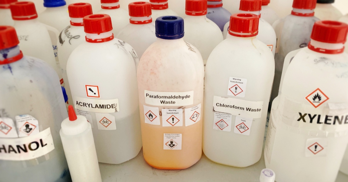 Assorted plastic bottles with blue or red caps are labeled with the various types of chemical waste they contain.