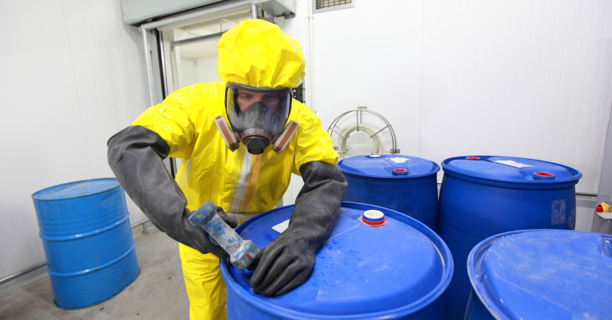 A man in yellow personal protective equipment and a respirator working with five blue hazmat barrels.