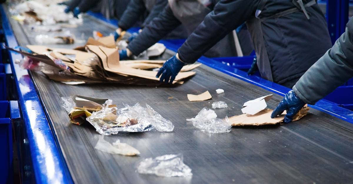 A close-up of the gloved hands of multiple workers sorting waste on a conveyor belt and removing cardboard.