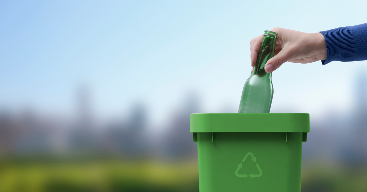 A hand drops a green glass bottle into a green, plastic bin with the recycling symbol on it outside.