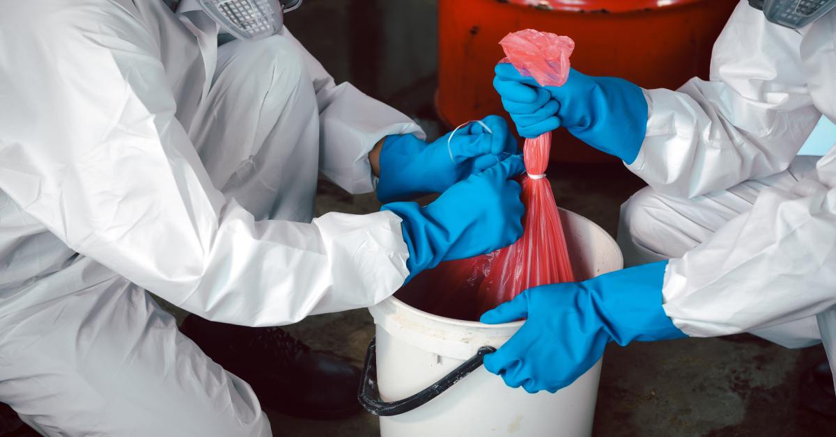 Two people in protective hazmat suits are preparing to dispose harmful chemicals and waste in a red bag.