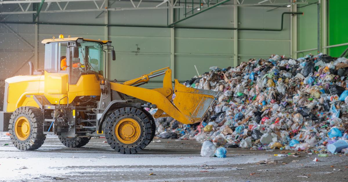 A yellow bulldozer in a recycling plant is sorting through waste piles in an industrial, empty room.