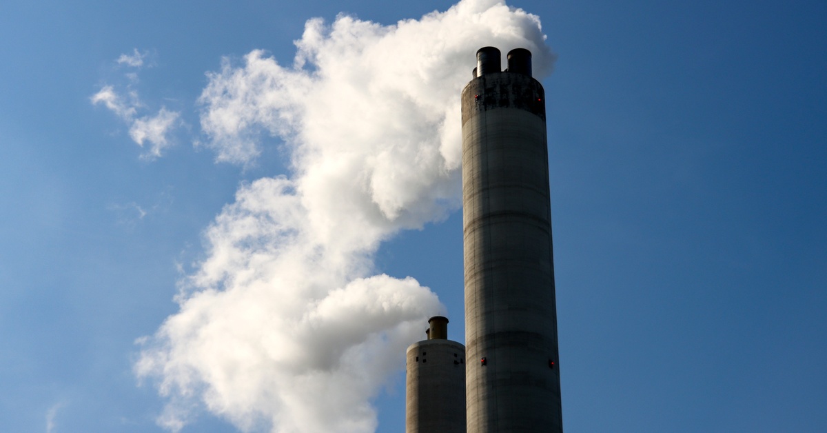 A close-up of two large industrial chimneys at a factory with steam and smoke coming out of them on a sunny day.