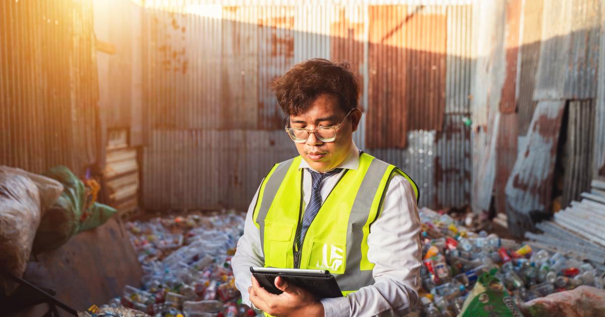 Man in reflective vest and tie checks a tablet while standing among piles of plastic bottles in a scrapyard.