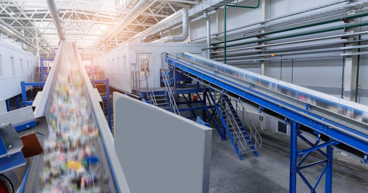 Conveyor belts carry mixed recyclable waste through a bright industrial sorting facility with metal beams overhead.