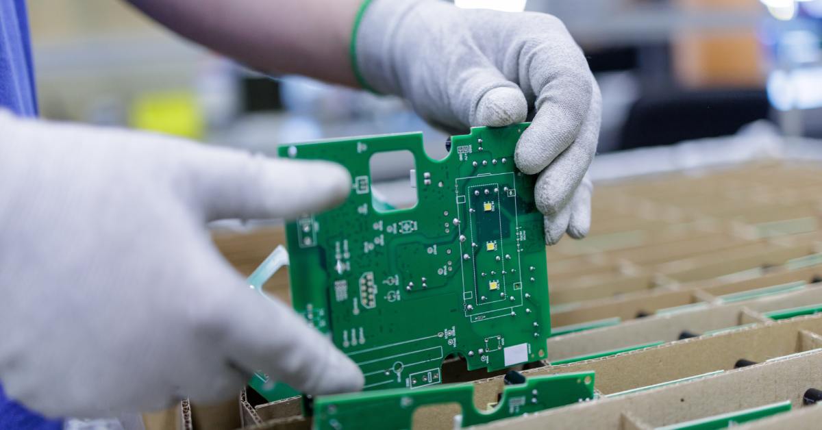 Gloved hands hold green circuit board over tray, inspecting electronic components during assembly process.