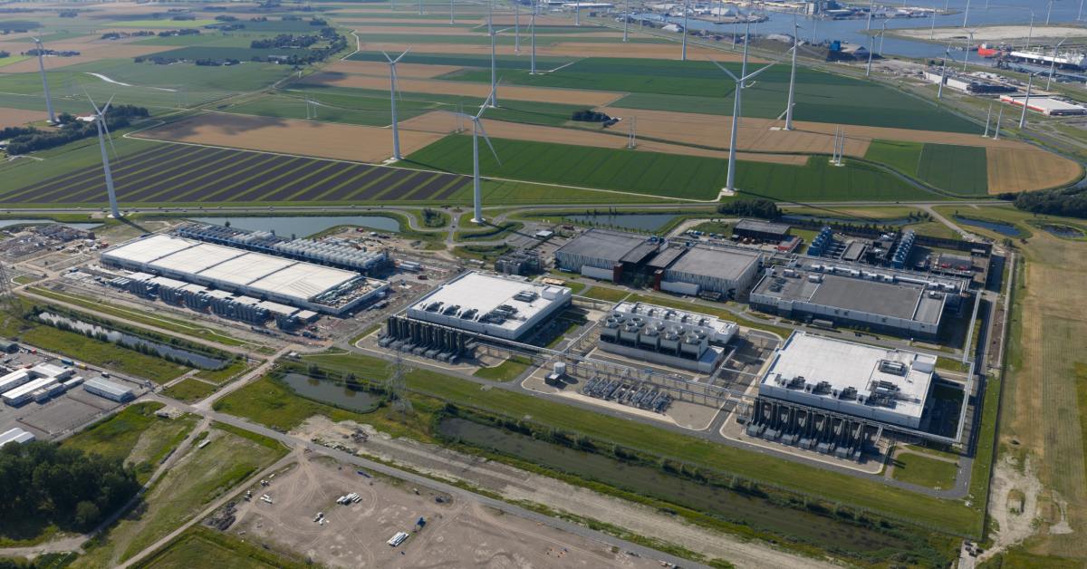 Aerial view of industrial complex near farmland with multiple wind turbines, roads, and water channels in distance.