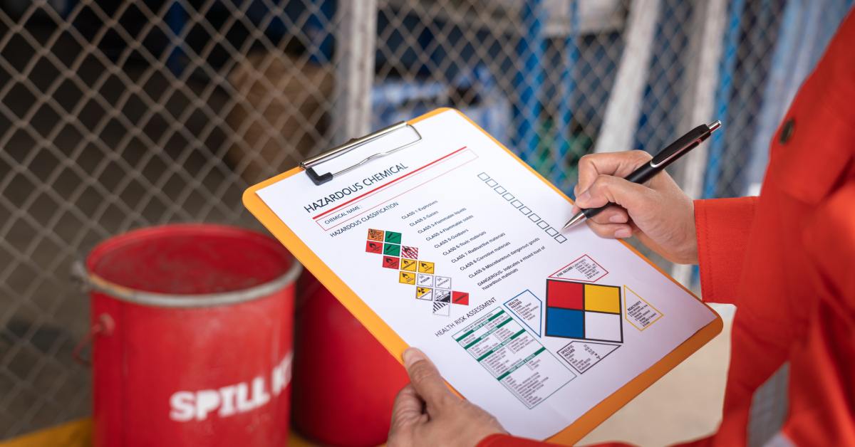 Worker writes on hazardous chemical form attached to clipboard, with warning symbols and barrels in background.