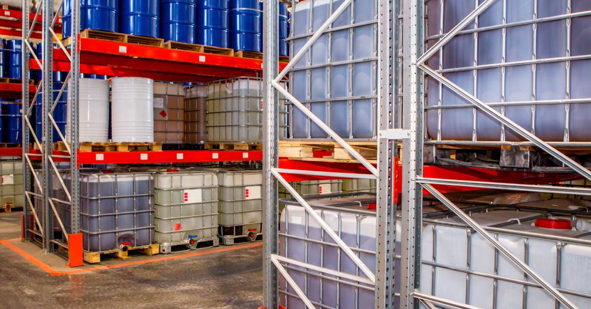 Industrial warehouse shelves storing large plastic containers and blue barrels filled with liquids on pallets.