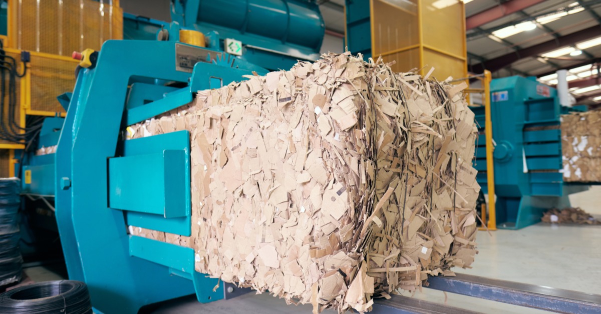 Large bale of shredded cardboard emerges from a blue recycling baler inside an industrial processing facility.