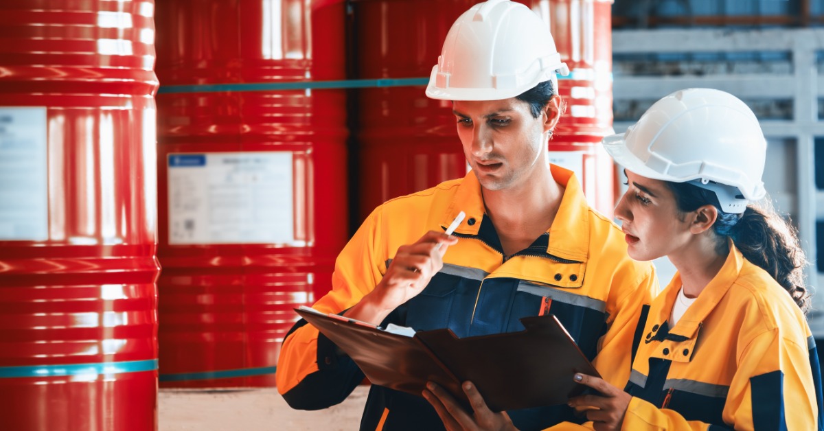Workers wearing white hard hats check documents beside stacked red barrels in an industrial storage area.