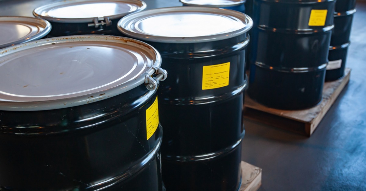 Black steel drums with sealed lids and yellow labels stored on wooden pallets in an industrial warehouse.