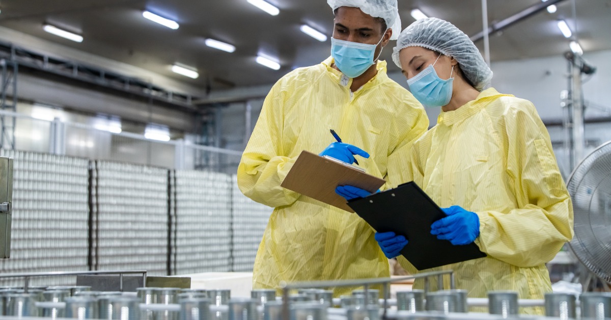  Manufacturing employees wearing PPE inspect paperwork on clipboards next to metal containers on a factory floor.