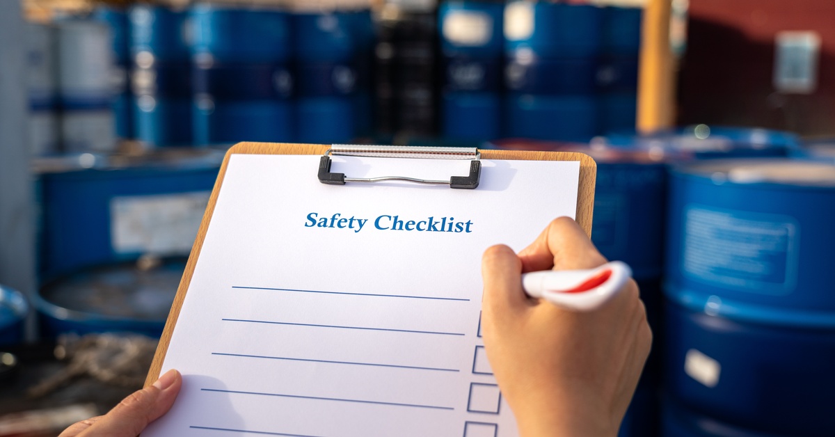 Workers in yellow safety uniforms and white hard hats examine documents near rows of large red industrial drums.