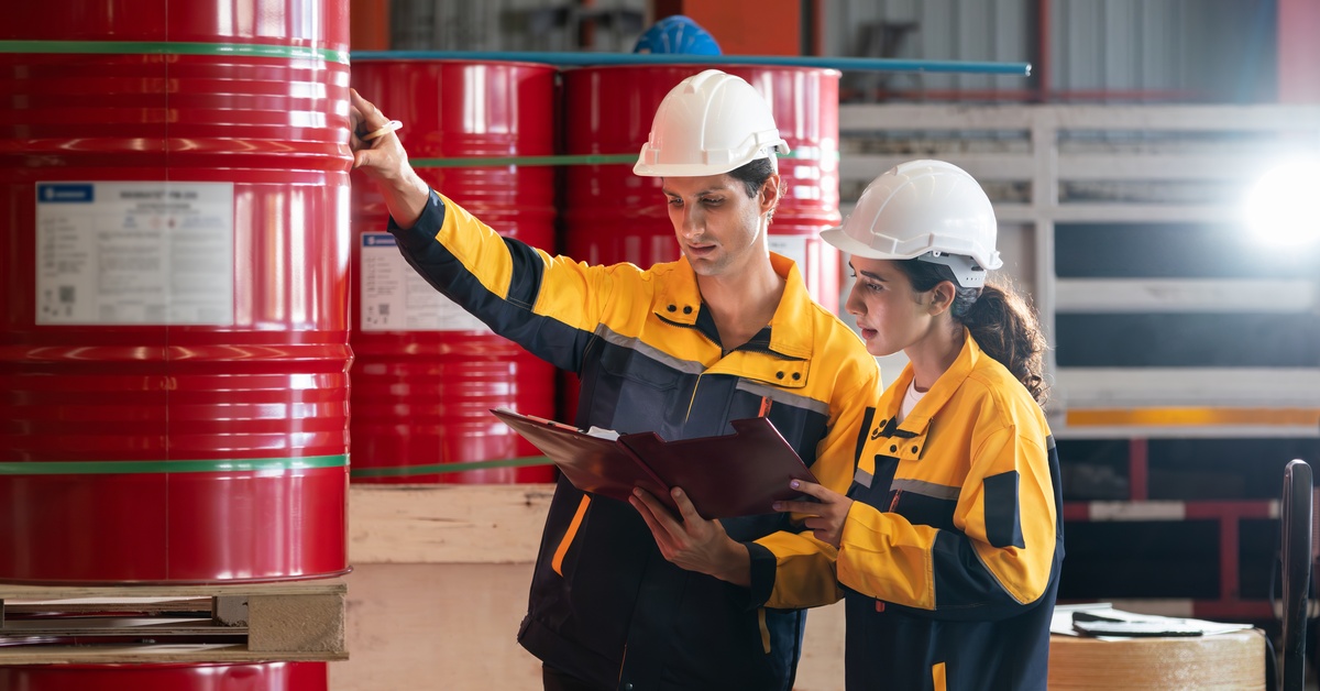 Workers in yellow safety uniforms and white hard hats examine documents near rows of large red industrial drums.