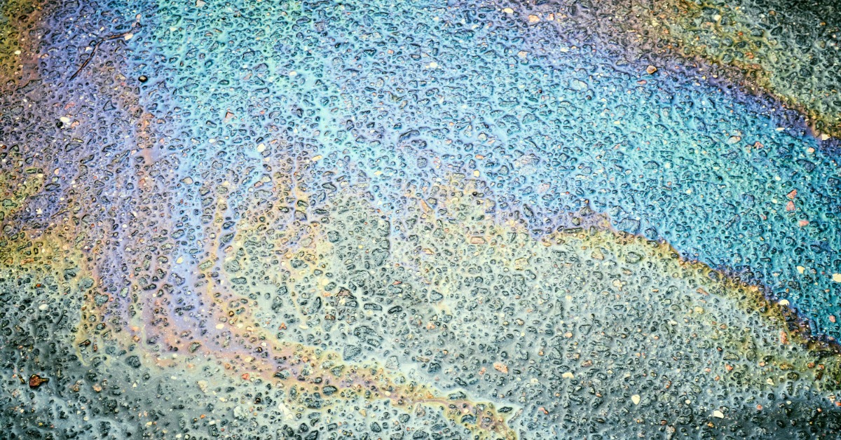 Rainbow-colored oil residue spreads across rough pavement, creating shimmering blue, green, and purple patterns.