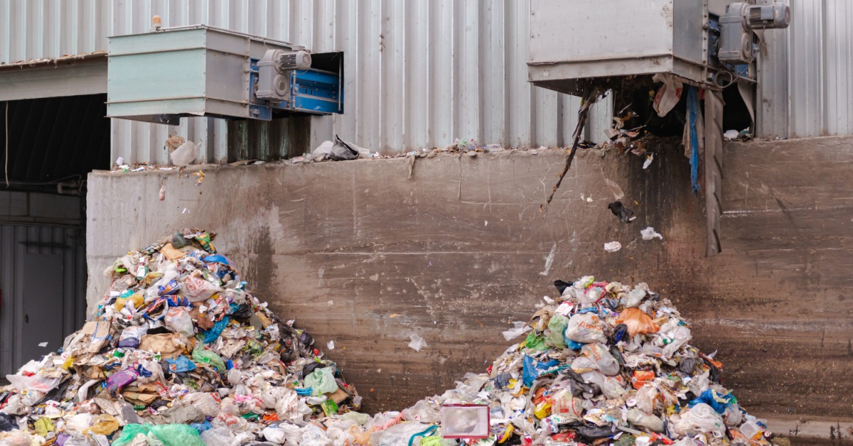 Industrial waste processing site with metal conveyor outlets depositing plastic bags, paper, and debris into trash piles.